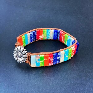 Pode incluir: Um bracelete colorido feito com contas de pedra multicoloridas e couro marrom. O bracelete tem um fecho de flor de prata.