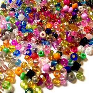 May include: A large assortment of colourful glass beads in various shapes and sizes. The beads are scattered on a white surface.
