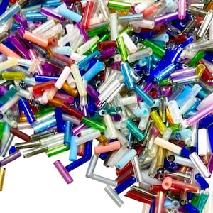 May include: A close-up of a collection of colourful glass seed beads. The beads are cylindrical and in various colours, including red, blue, green, pink, and clear. The beads are scattered against a white backdrop.