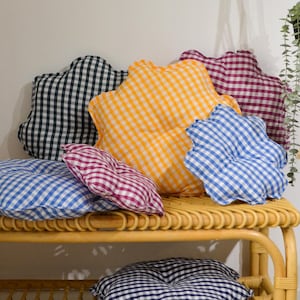 Può includere: Una collezione di cuscini a fantasia gingham in vari colori, tra cui blu, giallo, nero e bordeaux, disposti su una panca in rattan naturale. I cuscini hanno una forma smerlata unica. Una pianta sospesa è visibile sullo sfondo.