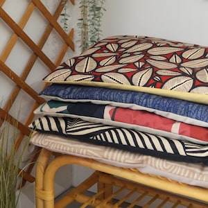 Peut inclure: Une pile de coussins décoratifs aux motifs et couleurs variés. Le coussin supérieur présente un fond rouge avec un motif de feuilles blanches et beiges. Les coussins sont empilés sur un banc en rotin brun clair.