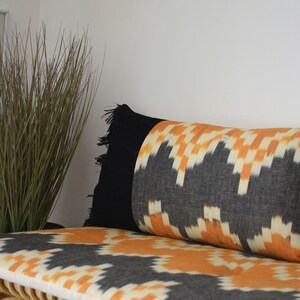 Peut inclure: Un coussin à motif noir et orange avec une frange noire posé sur un banc en bois clair avec un coussin assorti.