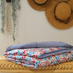 Peut inclure: Une couette pliée en tissu vichy bleu et blanc avec un imprimé floral sur l'autre face. La couette est posée sur un banc en osier.