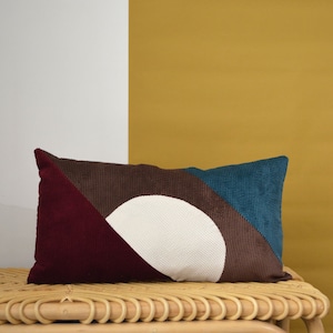 Peut inclure: Un coussin lombaire brun, bleu sarcelle et bordeaux avec un motif de cercle blanc. Le coussin est posé sur un banc en osier.