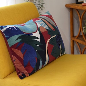 Op de afbeelding: Een gele fauteuil met een kleurrijk abstract patroon kussen. Het kussen heeft een mix van rode, blauwe, groene en witte kleuren.
