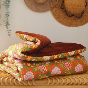 Peut inclure: Une couette en velours marron et à motifs floraux pliée sur un banc en osier tressé. La couette a un fond vert avec des motifs floraux orange, jaunes et blancs.