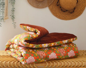 May include: A brown velvet and floral patterned quilt folded on a woven wicker bench. The quilt has a green background with orange, yellow, and white floral designs.