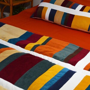 Op de afbeelding: Een gestreept corduroy bedspreid met een witte rand en een bijpassend kussen. De bedspreid is gemaakt van verschillende kleuren, waaronder oranje, geel, bruin, rood, blauw en groen.