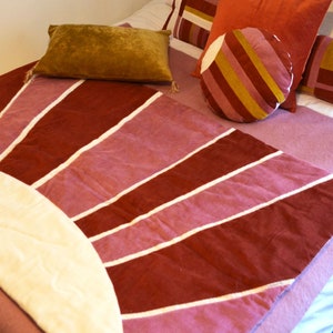 Può includere: Una coperta in velluto a coste rosa e rossa con un design a raggiera bianco. La coperta è su un letto con diversi cuscini, tra cui un cuscino rotondo a righe.