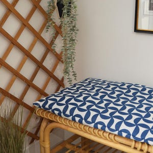 May include: A rattan bench with a blue and white geometric patterned cushion. The bench has a slatted shelf and curved legs. A hanging plant and wooden trellis are in the background.