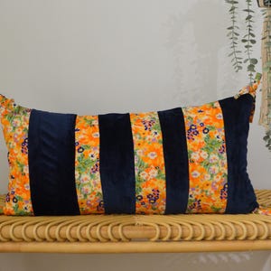 May include: Rectangular cushion with a floral pattern and navy blue velvet stripes. The cushion features a vibrant orange and yellow floral print with green leaves. The cushion is placed on a woven bench.