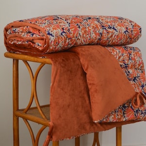Peut inclure: Une courtepointe pliée à motifs orange et bleu repose sur une table d'appoint en bambou. Un jeté en velours orange assorti est drapé sur le côté. L'ensemble crée une ambiance chaleureuse.