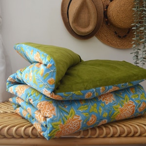 Könnte beinhalten: Eine gefaltete blaue und orangefarbene Steppdecke mit Blumenmuster und grünem Samtrücken. Die Steppdecke liegt auf einer geflochtenen Bank.