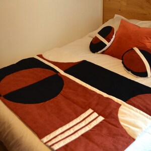 Peut inclure: Un lit avec une couette blanche, noire et rouille à motif géométrique. Il y a trois oreillers sur le lit, un est de couleur rouille et deux sont noirs et blancs.