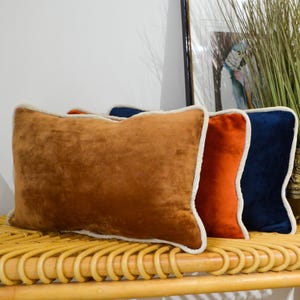 Puede incluir: Tres cojines rectangulares de terciopelo en tonos marrón, naranja y azul marino, cada uno con un ribete color crema. Los cojines están colocados sobre un banco tejido natural, creando un ambiente acogedor.