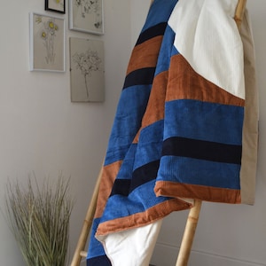Peut inclure: Une courtepointe rayée bleue, marron et blanche avec un motif géométrique. La courtepointe est pliée et drapée sur une échelle en bois.