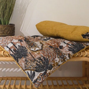 May include: A patterned cushion with a brown, black and white design sits on a woven wicker bench. A solid mustard yellow cushion is placed on top of the patterned cushion.