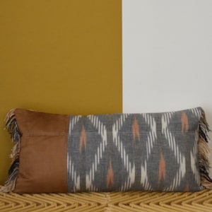 May include: Rectangular cushion featuring a brown leather section and a grey and white patterned fabric. The cushion has a fringe trim. Ideal for home decor, it measures approximately 40 cm in length.
