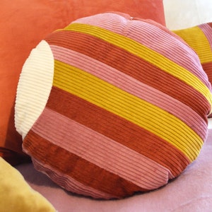 Può includere: Un cuscino rotondo a righe con un cerchio bianco al centro. Il cuscino è realizzato in tessuto di velluto a coste e presenta strisce rosa, rosse e gialle.
