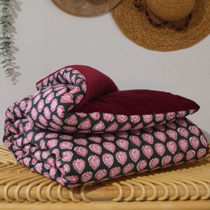 Könnte beinhalten: Eine gefaltete Bettdecke mit einem dunkelgrünen und rosa Paisley-Muster, kombiniert mit einem einfarbigen bordeauxfarbenen Kissen. Die Bettdecke liegt auf einer hellbraunen, gewebten Oberfläche, mit Strohhüten im Hintergrund.