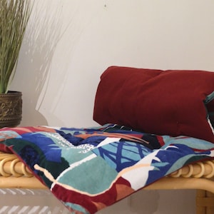 May include: A colorful patterned cushion with a burgundy velvet backing. The cushion is rolled up and placed on a wicker bench.