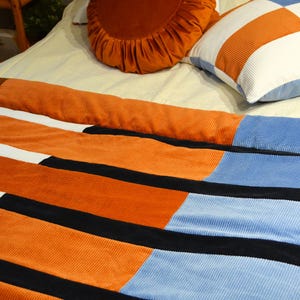 Könnte beinhalten: Eine Cord-Bettdecke mit einem Patchwork-Design in Orange, Weiß, Schwarz und Hellblau. Zwei Kissen sind ebenfalls zu sehen: ein rundes orangefarbenes Kissen und ein quadratisches Kissen mit einem orange-weiß karierten Muster. Das Ganze liegt auf einem Bett.