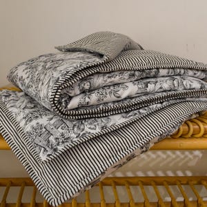 Op de afbeelding: Een stapel zwart-wit patronen quilts. De bovenste quilt heeft een zwart-wit gestreept patroon op de achterkant. De quilts zijn gestapeld op een natuurlijke rieten plank. De quilts hebben een toile-patroon.