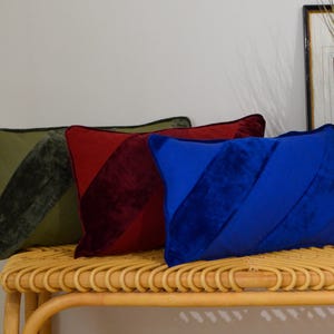 Peut inclure: Trois coussins rectangulaires en velours vert olive, rouge et bleu, avec une rayure diagonale. Ils sont disposés sur un banc en osier brun clair. Une image encadrée est visible en arrière-plan.