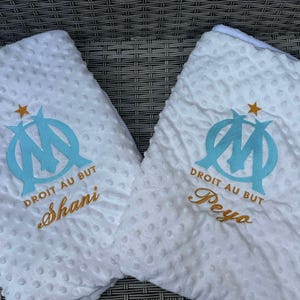 Peut inclure: Deux couvertures blanches pour bébé, texturées. Chacune présente un logo brodé bleu clair et le texte "DROIT AU BUT". L'une porte le nom "Shani" brodé en or, l'autre "Peyo".