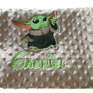 Peut inclure: Une couverture de bébé beige avec une image brodée verte d'un personnage de la franchise Star Wars, connu sous le nom de "Baby Yoda", et le nom "Gabriel" brodé sous l'image.