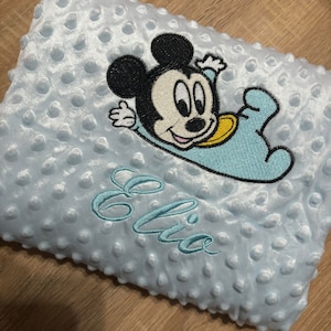 Peut inclure: Une couverture de bébé bleu clair avec une surface texturée à pois. La couverture présente une image brodée de Mickey Mouse dans une tenue de bébé. Le nom "Elia" est brodé en script bleu clair assorti.