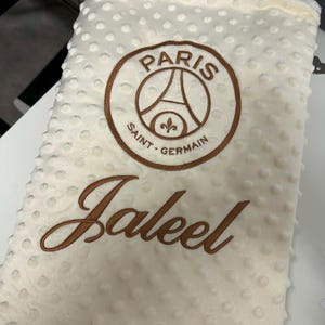 May include: Cream-colored baby blanket with a textured, dotted surface. Features an embroidered brown Paris Saint-Germain logo and the name "Jaleel" in brown script. A soft and cozy blanket.