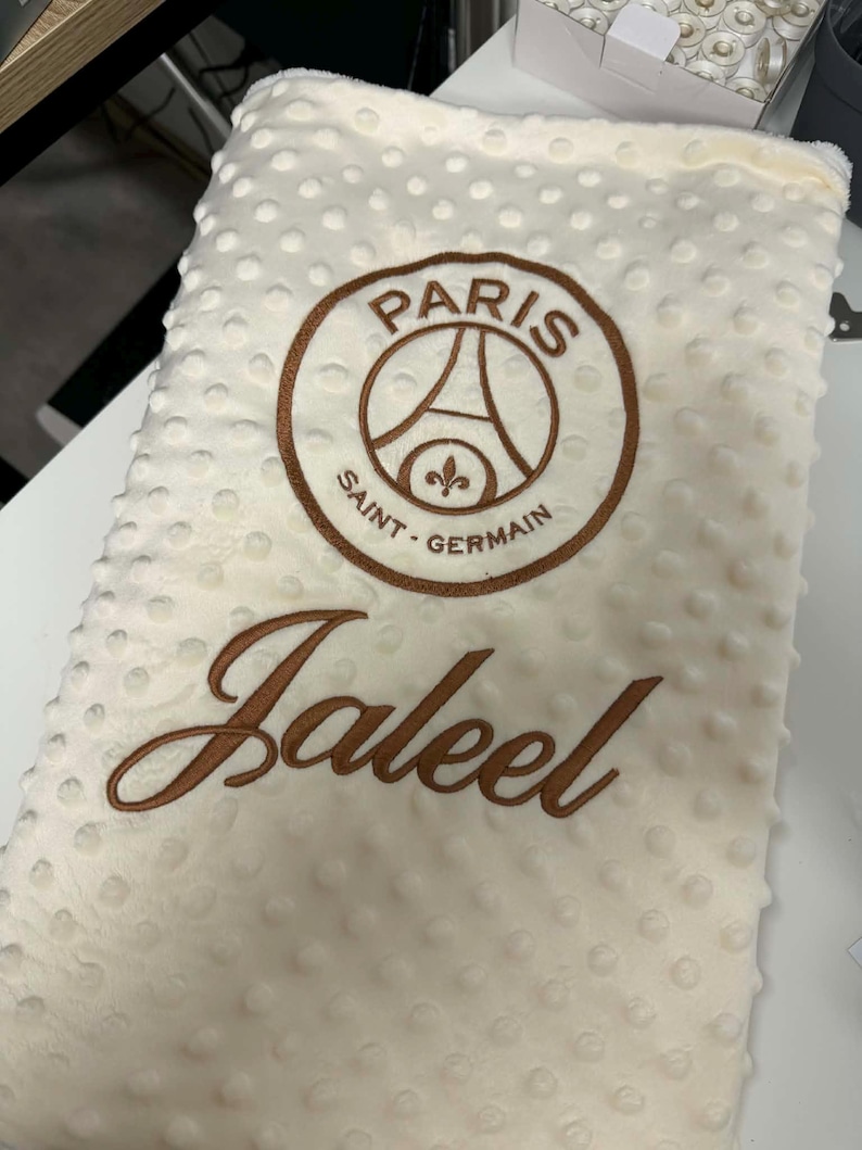 May include: Cream-colored baby blanket with a textured, dotted surface. Features an embroidered brown Paris Saint-Germain logo and the name "Jaleel" in brown script. A soft and cozy blanket.