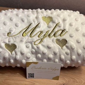 Peut inclure: Une couverture blanche pour bébé avec un nom brodé en or "Myla" et trois cœurs dorés. La couverture a une surface texturée et bouffante.