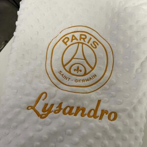 May include: White, textured blanket with a gold embroidered Paris Saint-Germain logo and the name "Lysandro" in gold script. The blanket has a soft, dotted pattern.