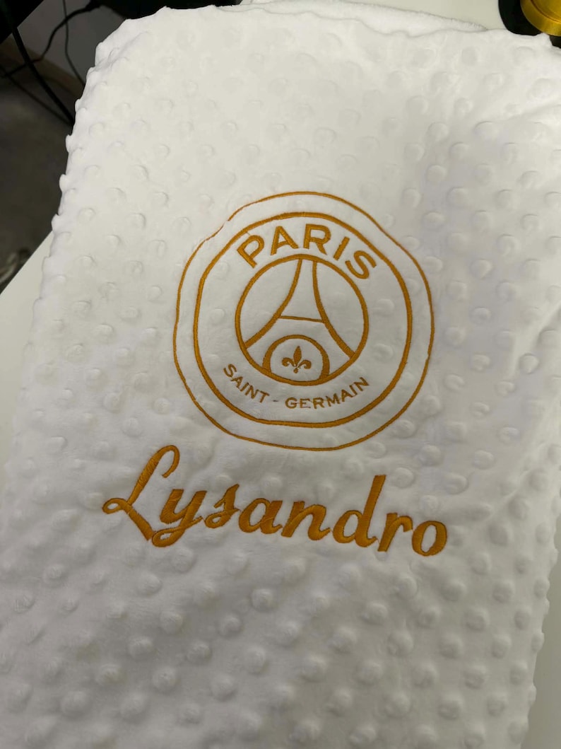 May include: White, textured blanket with a gold embroidered Paris Saint-Germain logo and the name "Lysandro" in gold script. The blanket has a soft, dotted pattern.