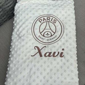 May include: White baby blanket with a textured, dotted surface. Features the Paris Saint-Germain logo in brown, along with the name "Xavi" in a matching brown script. The blanket is folded.