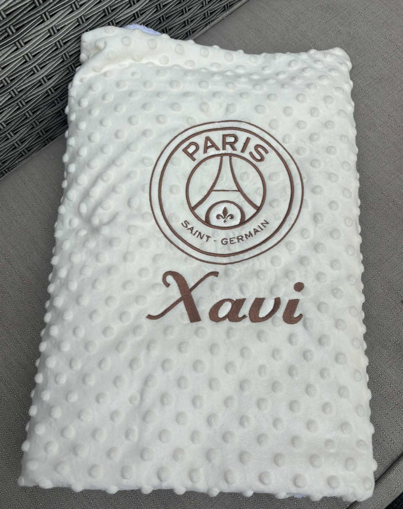 May include: White baby blanket with a textured, dotted surface. Features the Paris Saint-Germain logo in brown, along with the name "Xavi" in a matching brown script. The blanket is folded.