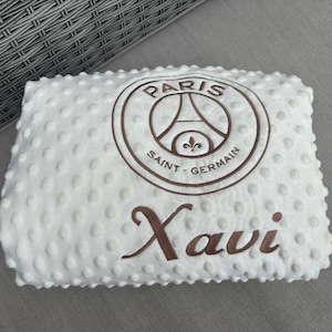 May include: White baby blanket with a textured surface and brown embroidered details. Features the Paris Saint-Germain logo and the name "Xavi". The blanket is rectangular and appears soft.