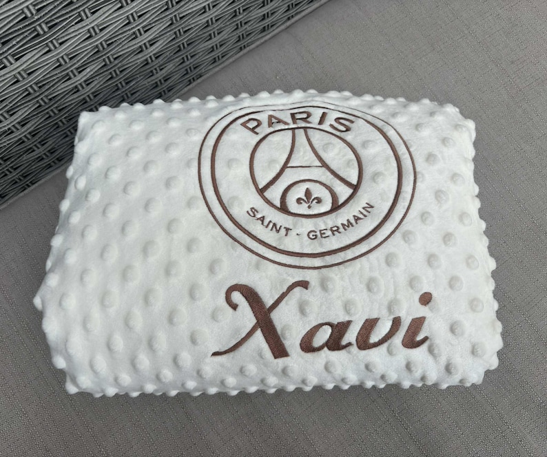 May include: White baby blanket with a textured surface and brown embroidered details. Features the Paris Saint-Germain logo and the name "Xavi". The blanket is rectangular and appears soft.