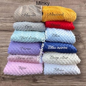 May include: A stack of Minky fabric swatches in various colors, including gray, yellow, red, blue, and pink. Each swatch is labeled with its color name in cursive script. The fabric has a textured, dotted surface.