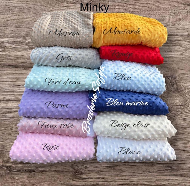 May include: A stack of Minky fabric swatches in various colors, including gray, yellow, red, blue, and pink. Each swatch is labeled with its color name in cursive script. The fabric has a textured, dotted surface.