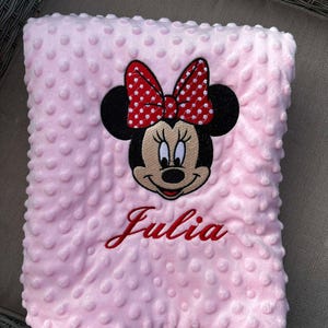 Peut inclure: Une couverture rose pour bébé avec une broderie de Minnie Mouse à pois rouges et blancs et le nom "Julia" brodé en rouge.