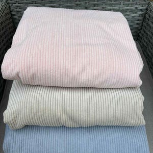 May include: Three folded corduroy fabric pieces in pastel colors: light pink, light gray, and light blue. The fabric has a ribbed texture, and the pieces are stacked on top of each other. The image is taken from a slightly elevated angle.
