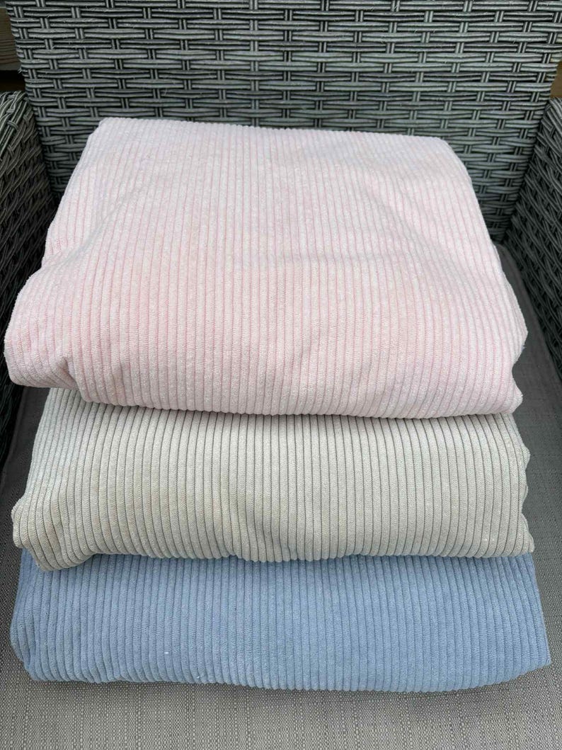 May include: Three folded corduroy fabric pieces in pastel colors: light pink, light gray, and light blue. The fabric has a ribbed texture, and the pieces are stacked on top of each other. The image is taken from a slightly elevated angle.