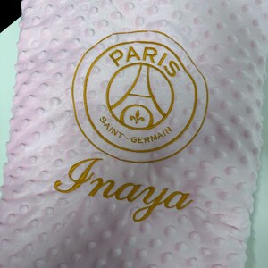 May include: Pink baby blanket with a textured surface and a gold embroidered Paris Saint-Germain logo and the name "Inaya". The blanket is a soft, plush material.