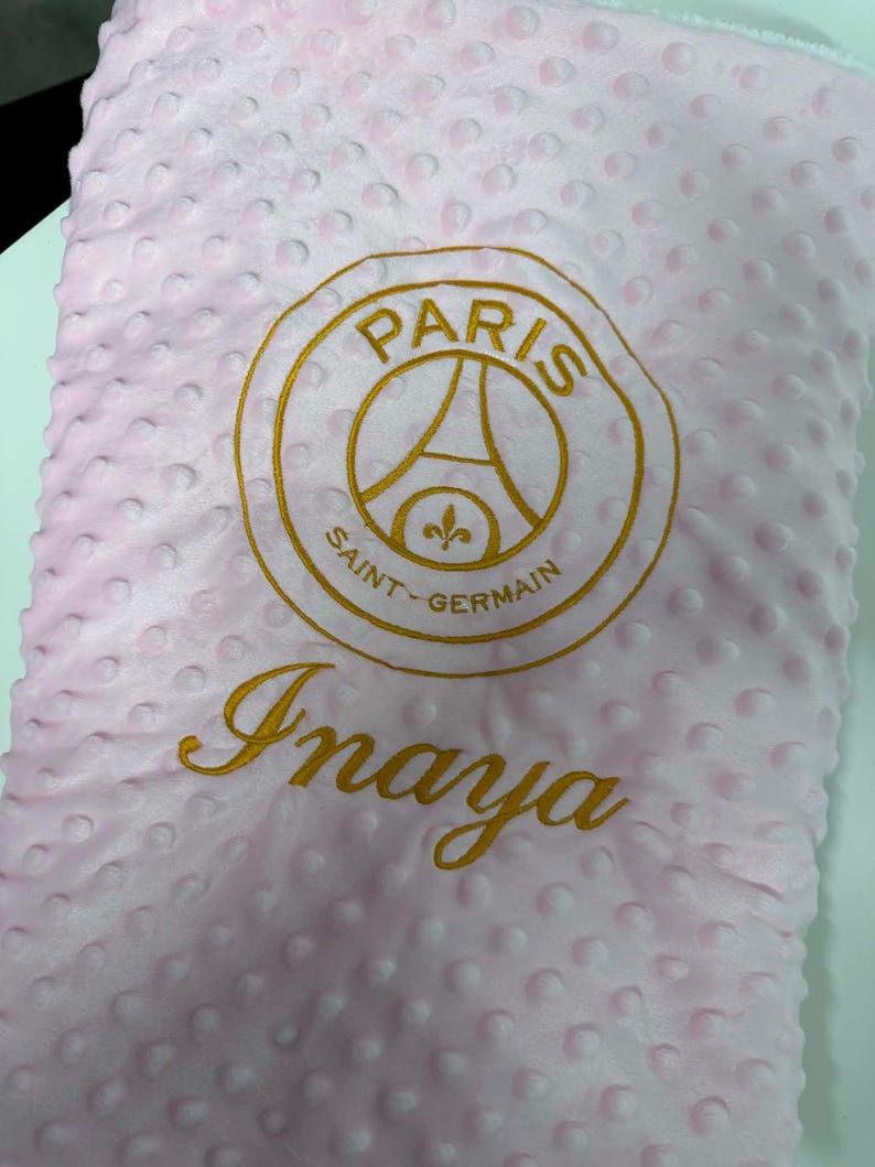 May include: Pink baby blanket with a textured surface and a gold embroidered Paris Saint-Germain logo and the name "Inaya". The blanket is a soft, plush material.