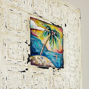 May include: A white framed picture with a distressed, aged look. The picture features a colorful painting of a palm tree on a beach.