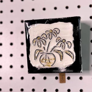 May include: A small black square box with a white background and a gold and black drawing of flowers in a vase. The flowers are stylised and have a simple design.