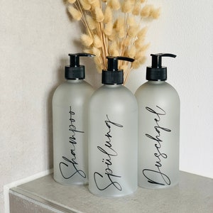 Könnte beinhalten: Drei milchglasartige Pumpflaschen mit schwarzen Pumpen und Etiketten. Die Etiketten sind in schwarzer Schrift geschrieben und sagen "Shampoo", "Spülung" und "Duschgel".